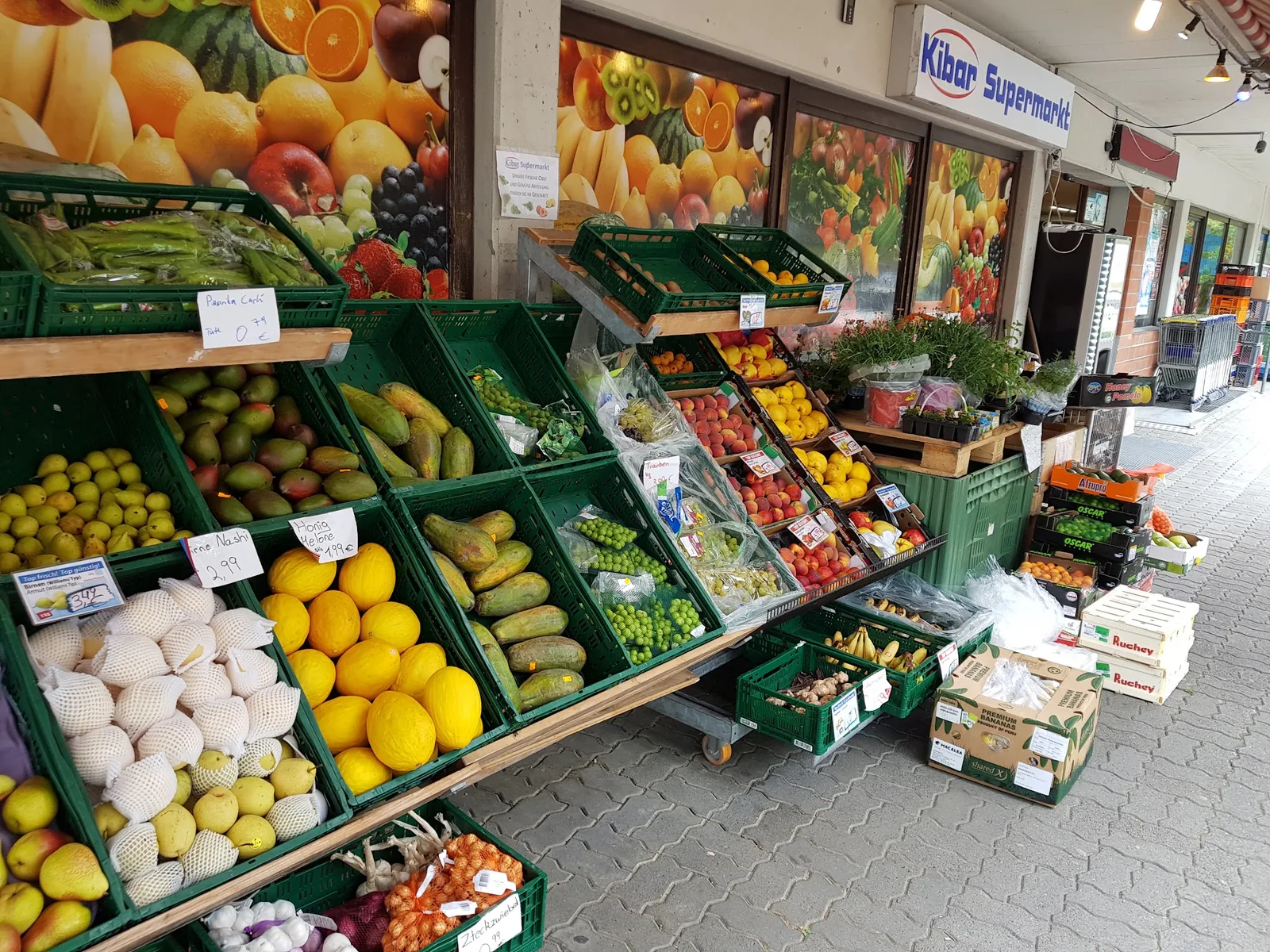 Obst- und Gemüseauslage in Holzsteigen vor dem Laden, darüber das Firmenschild Kibar Supermarkt