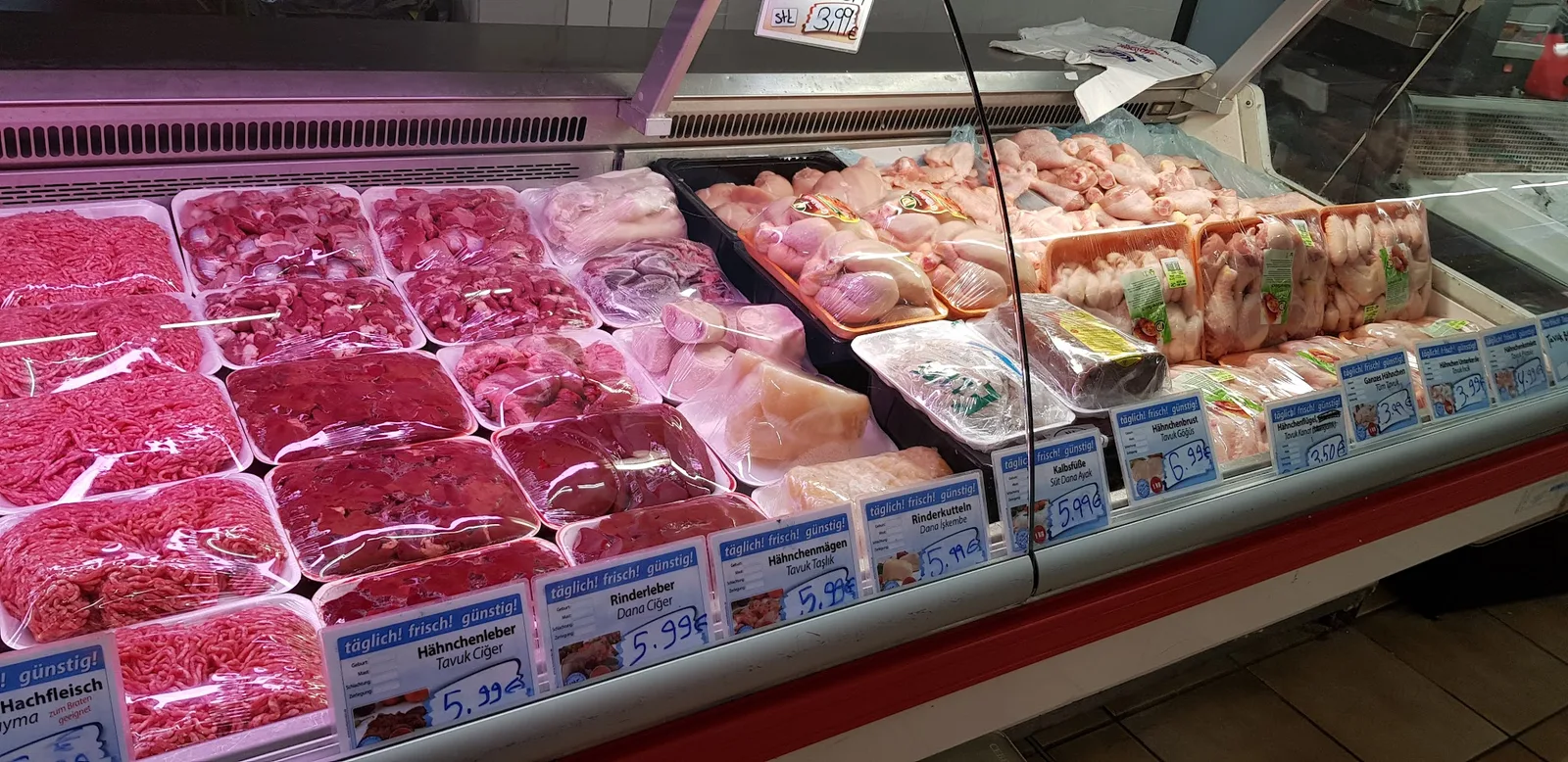 Bedientheke in ganzer Breite mit Hackfleisch, Innereien und Hähnchenteilen, jedes Schild zweisprachig beschriftet